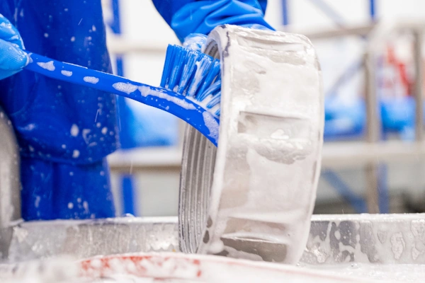 An Ecowize worker in blue protective clothing cleaning a stainless steel can with a blue brush and soapy detergent in a food processing facility.