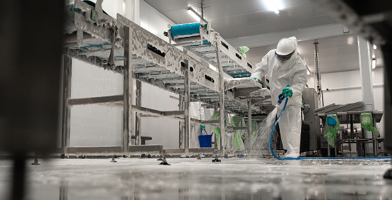 Food processing sanitation technician pressure washing conveyor equipment in a production facility while wearing full protective clothing, demonstrating hygienic cleaning and food safety compliance.